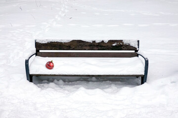 Forgotten red Christmas ball with painted snowflakes lying on snowy empty brown wooden bench outdoor in the park in winter day front view