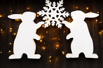 Two white styrofoam bunny figures holding snowflake decoration with yellow string lights on dark wooden wall.