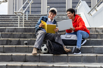 Diverse male student friends discussing study notes on campus stairs with laptop beside soccer ball