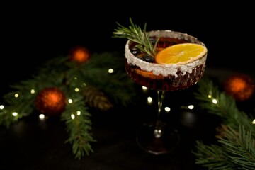 Festive Christmas cocktail served in an elegant glass with orange slice, blueberries and rosemary garnish. Surrounded by holiday lights, pine branches and Christmas ornaments on a dark background. 