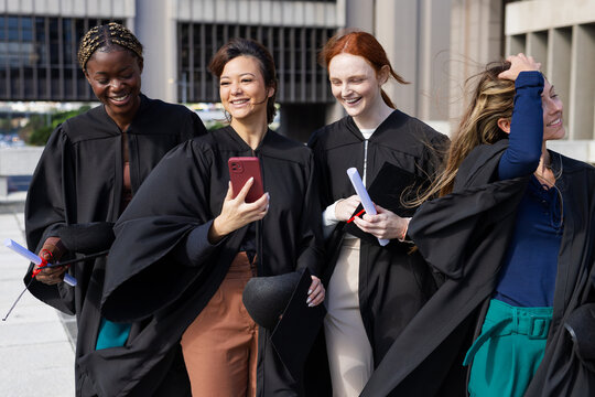Diverse female graduates walking along waterfront promenade carrying diplomas and smartphone