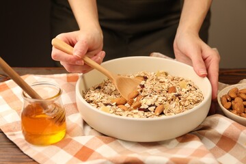 Woman making granola with oat flakes, dried fruits, almond nuts and honey at wooden table against grey background, closeup