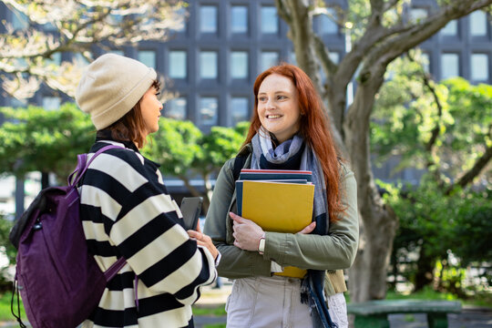 Diverse female students chatting on campus wearing backpack holding notebooks smartphone by benches