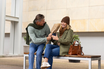 Couple sitting on bench at plaza, exchanging bouquet while woman scrolling smartphone
