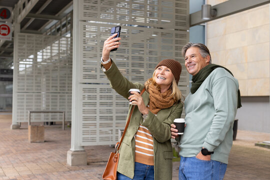 Senior couple holding coffee cups and taking selfie with smartphone on platform at bus stop sign