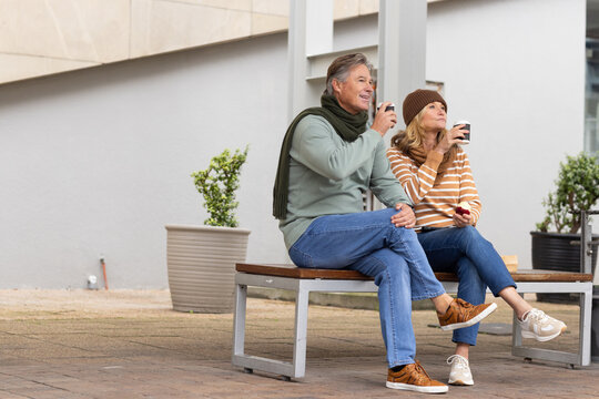 Senior couple sitting holding coffee cups on wooden bench in courtyard wearing scarf and beanie - Powered by Adobe