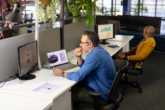 Diverse male coworkers collaborating on data charts using laptop amid plants in modern office
