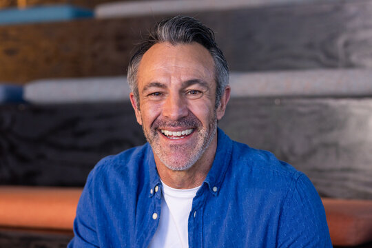 Mature adult man smiling while sitting on wooden bench steps in gym bleachers wearing blue shirt