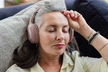 Mature adult woman reclining on dark sofa wearing pink headphones and relaxing in living room