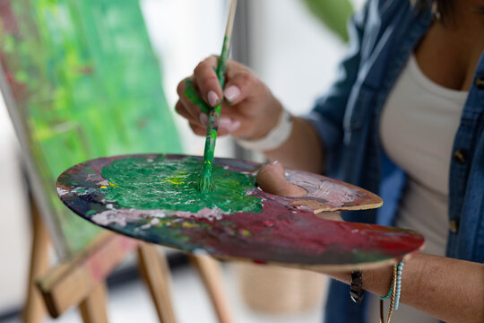 Woman wearing denim shirt mixing green paint on wooden palette while painting canvas at art studio