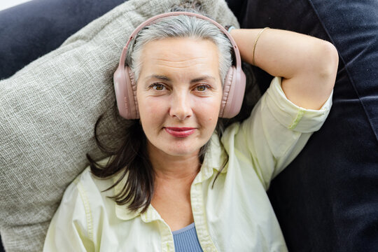 Senior woman reclining on sofa at home resting head on gray cushion and wearing gold headphones
