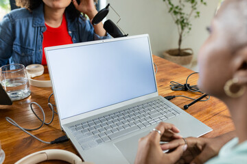 Diverse female coworkers recording podcast at home office table with laptop, microphone, headphones