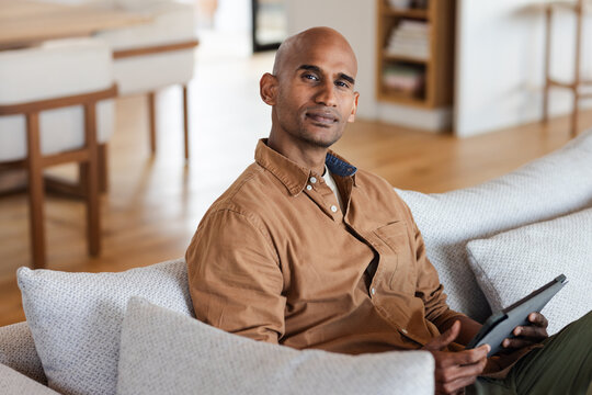 Asian man sitting on light-colored sofa holding tablet in modern home living room beside bookshelf