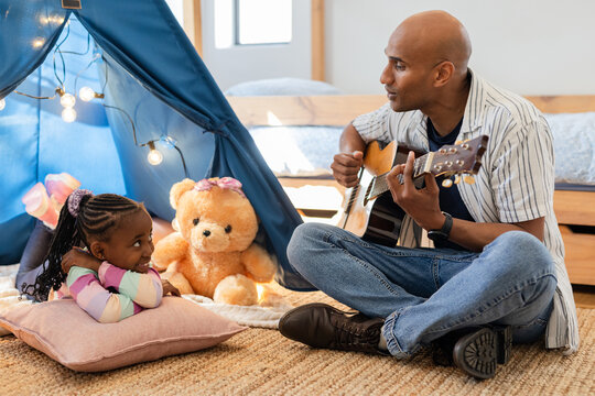 Diverse father and daughter sharing guitar lesson beside tent with teddy in bedroom, copy space