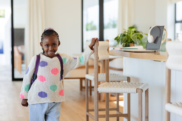 Smiling African American girl carrying backpack standing bar stool in bright home space, copy space