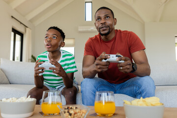African American father and son gripping controllers on sofa at home snacking on popcorn, juice