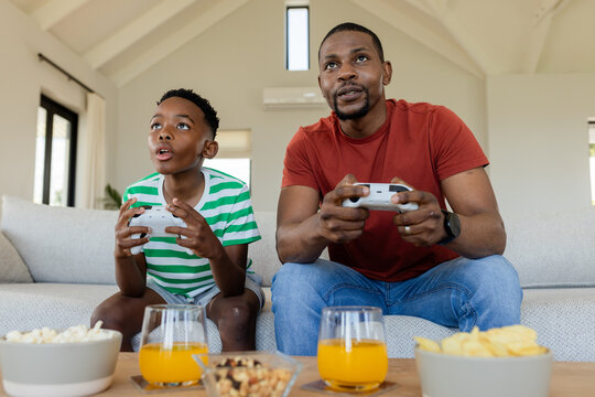 African American father and son gripping controllers on sofa at home snacking on popcorn, juice