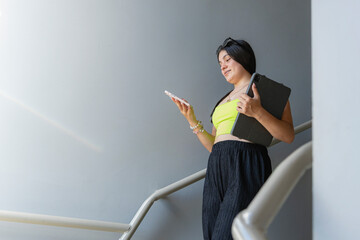 Latina teenager using smartphone while walking downstairs holding tablet