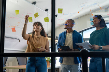 Diverse coworkers collaborating at office, using glass wall with sticky notes, marker and tablet