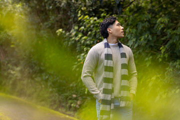 Hispanic man standing on mossy trail wearing knit sweater, striped scarf, blue jeans, gazing upward