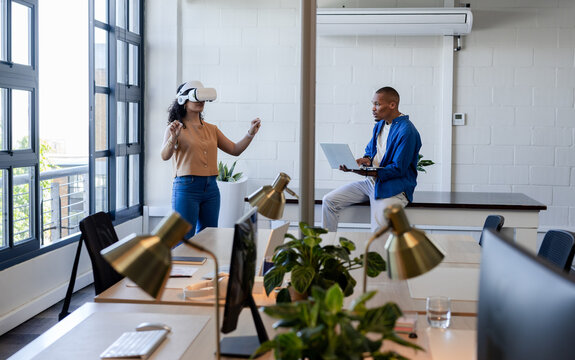 African American coworkers showing VR headset demo while using laptop in casual office with windows