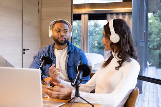 Diverse podcast hosts recording show at wooden table in paneled room with microphones, headphones