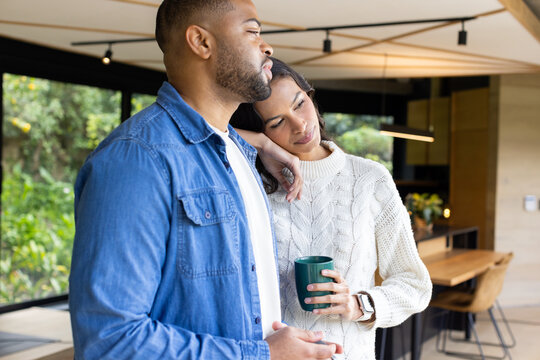 African American couple standing next to large window in open-plan kitchen holding green coffee mug - Powered by Adobe