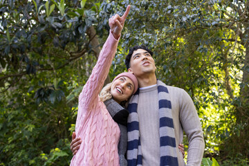 Diverse couple wearing pink beanie and striped scarf leaning under leafy canopy and pointing upward