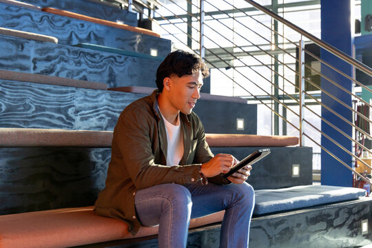 Asian man in mid-20s sitting on tiered bench in office lobby holding tablet computer