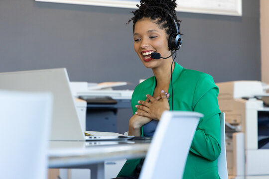 African American woman in green blazer smiling while touching chest at desk with laptop and headset