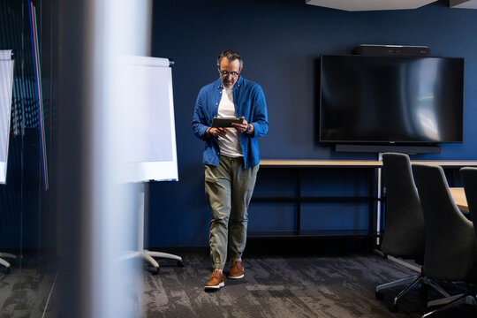 Middle-aged man walking across meeting room holding tablet, blank flip chart and chairs at table