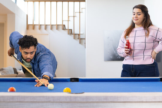 Couple leaning over blue pool table aiming cue stick at home rec room, holding bottled drink