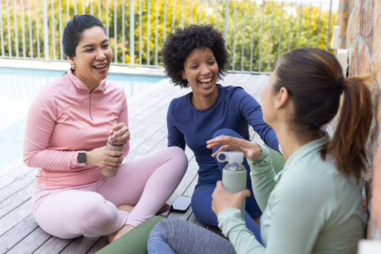 Diverse female friends sitting on yoga mats on pool deck in activewear laughing with water bottles - Powered by Adobe
