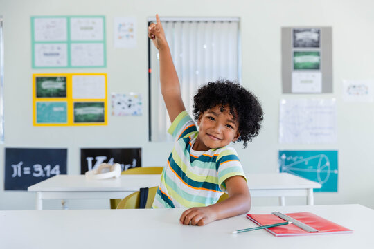 Male child sitting at desk in classroom wearing striped shirt raising hand near red notebook