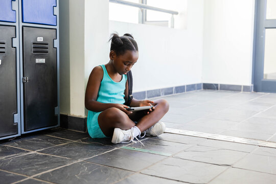 African American girl sitting on school hall using tablet in turquoise dress sneakers by lockers