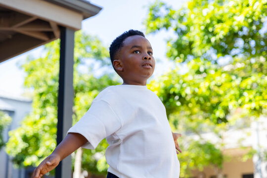 Male child balancing on porch near post and sunlit trees, wearing white tee and dark pants