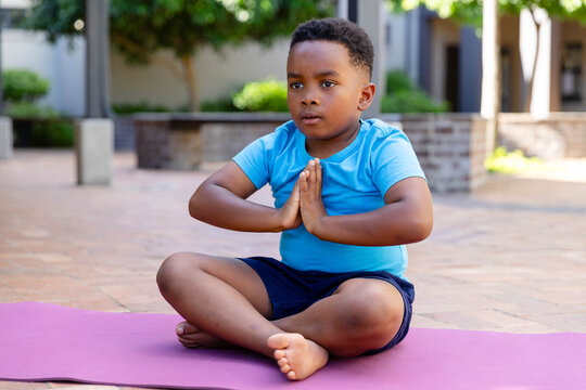 African boy sitting cross-legged on pink yoga mat in courtyard in light blue shirt pressing palms