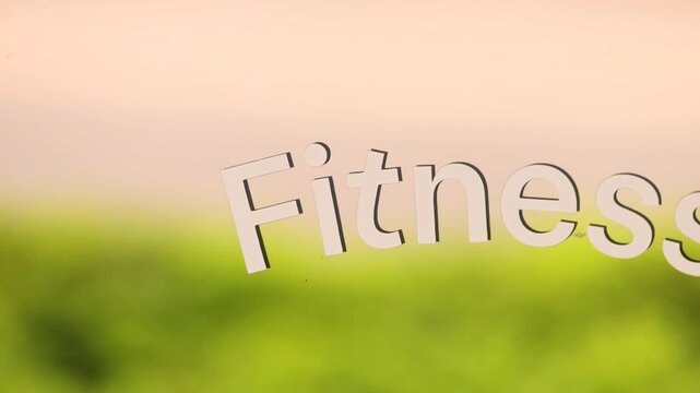 The word FITNESS is applied on transparent glass, creating a logo effect. Behind the glass, bright green grass reflects softly in bokeh, making the white letters stand out for modern design use.