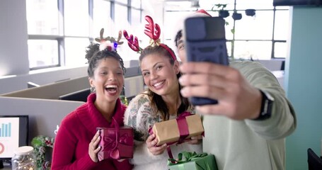 Diverse coworkers reaching for smartphone and capturing holiday selfie with gift boxes at office - Powered by Adobe