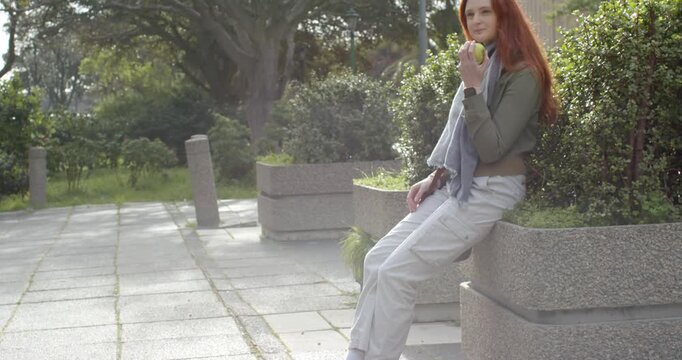 Receiving call, red-haired woman sitting on park bench lowering phone and biting apple, copy space