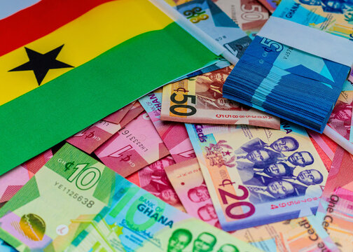 A collection of various Ghana Cedi banknotes scattered with the Ghanaian national flag and a banded stack of cash. A conceptual image for national economy, wealth, and banking in West Africa.