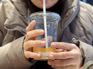 Hands Holding Iced Coffee Cup CloseUp, Student Gripping Cold Beverage With Both Hands At Cafe Table, Straw Near Lips, Condensation Beading On Plastic Cup, Casual Winter Jacket, Smartphone