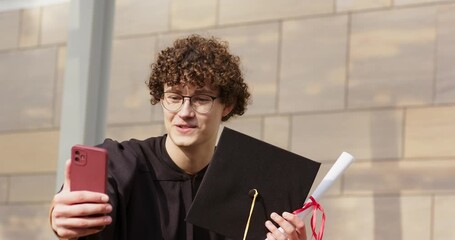 Male student extending red phone for live call, holding mortarboard and diploma by stone column