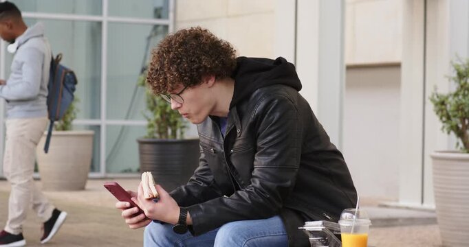 Male unwrapping sandwich, biting and scrolling phone on bench outside as passerby checking phone