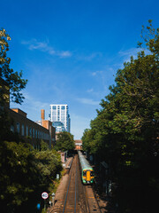 Passenger train traveling along railway tracks toward a modern cityscape framed by trees and bright blue sky, capturing dynamic urban transport scenery
