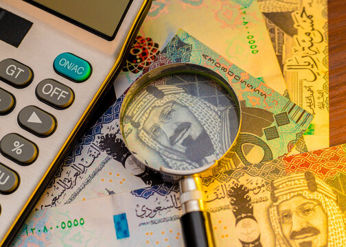 Close-up of Saudi Riyal banknotes featuring King Salman. A magnifying glass and calculator rest on the cash, symbolizing financial analysis, banking, and the Saudi Arabian economy.