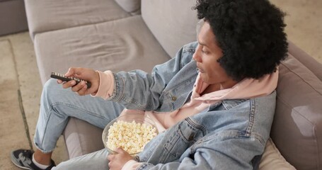 African American man clicking remote and munching popcorn on gray couch in living room, copy space