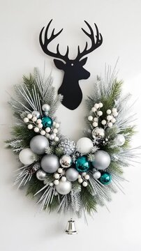 A stunning modern Christmas wreath with frosted pine, silver and teal ornaments, and white berries hangs beneath a bold black deer silhouette on a white wall. A delicate silver bell dangles below.