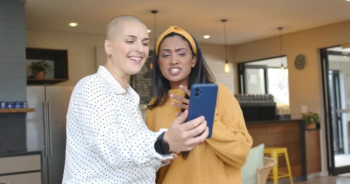 Diverse female friends holding blue smartphone, recording lip-sync while gesturing cup in cafe
