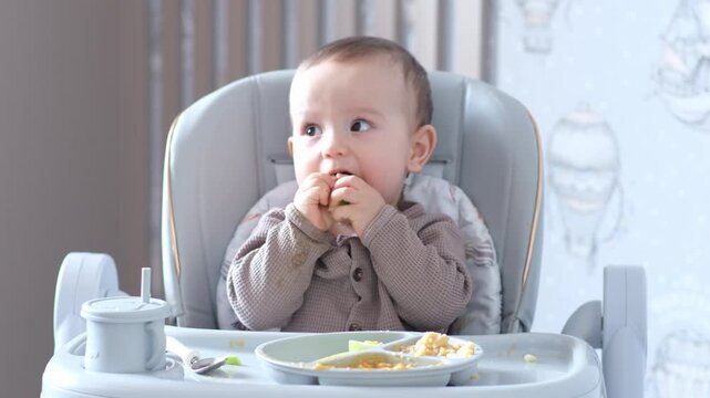 Baby in Highchair tastes Spoon Puree, Exploring Curious Infant ,Tests Utensils While Caregiver Guides Feeding, Hands Reach Tray, Mashed Vegetable On Plate, Soft